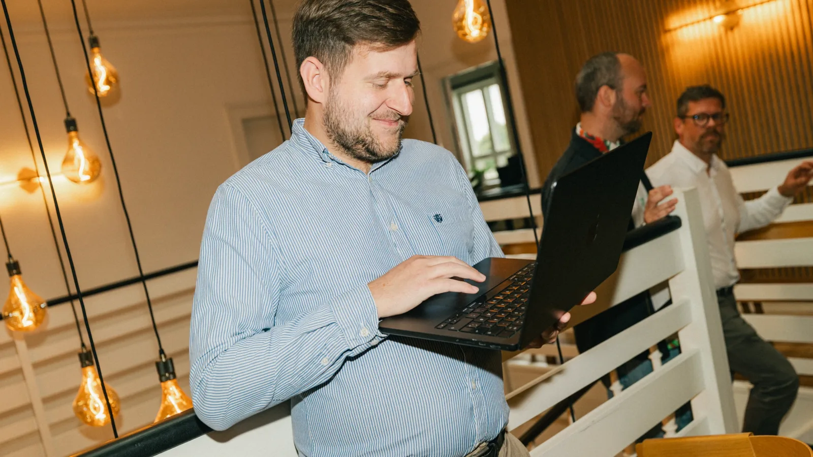 Smilende medarbejder i stribet skjorte arbejder på en bærbar computer på en trappe, mens to kolleger står i baggrunden