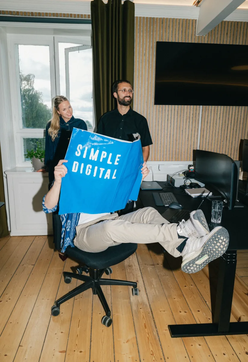 To kolleger står bag en medarbejder, der viser en blå Simple Digital-plakat frem