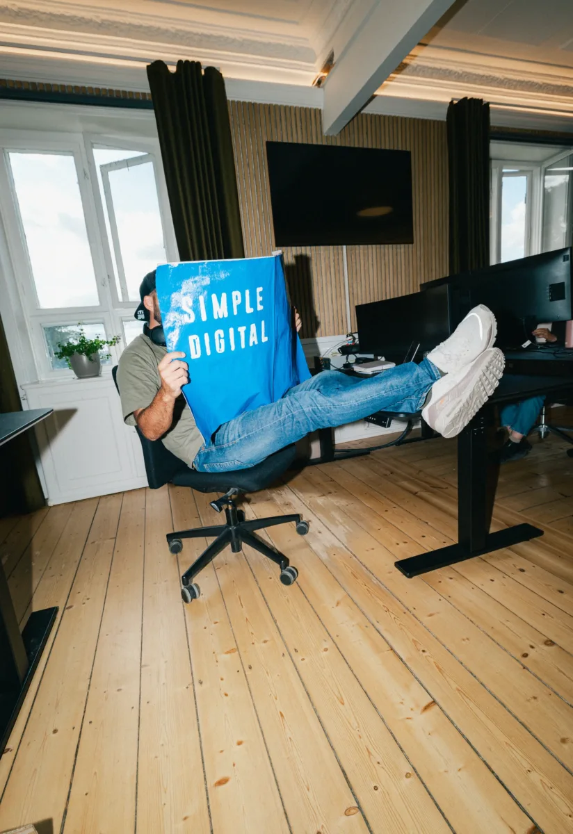 Medarbejder holder en Simple Digital-plakat op foran ansigtet med benene på bordet