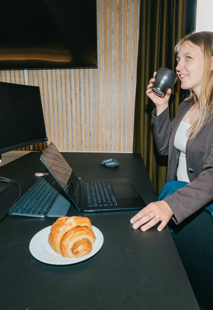 Medarbejder drikker af en kop ved et hæve-sænke-bord med croissant og bærbar computer