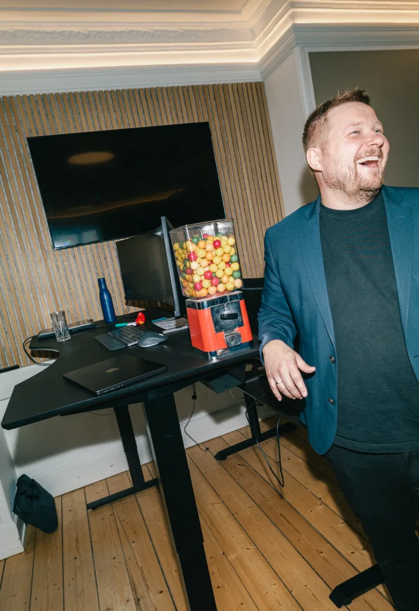 Medarbejder i blå blazer ler stående ved et skrivebord med en tyggegummiautomat