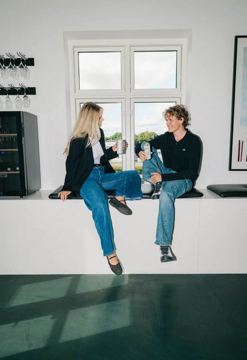 To kolleger sidder i en vindueskarm med hver sin dåse og taler sammen