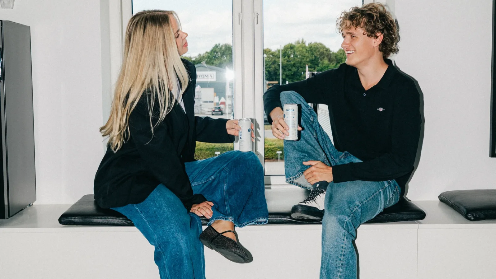 To kolleger sidder i en vindueskarm med dåser og kigger på hinanden