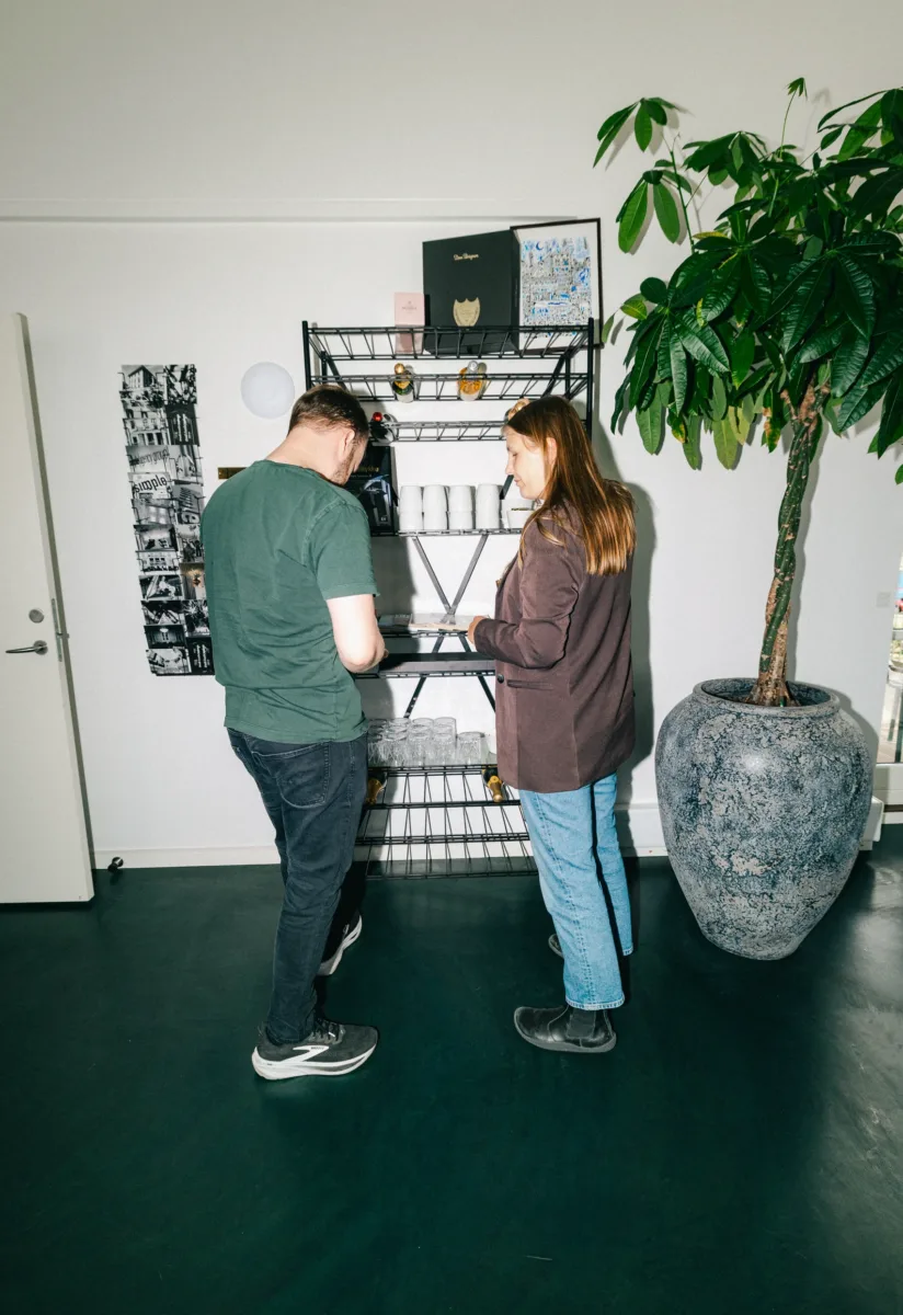 To kolleger står ved en trådreol med glas og flasker i et lyst lokale