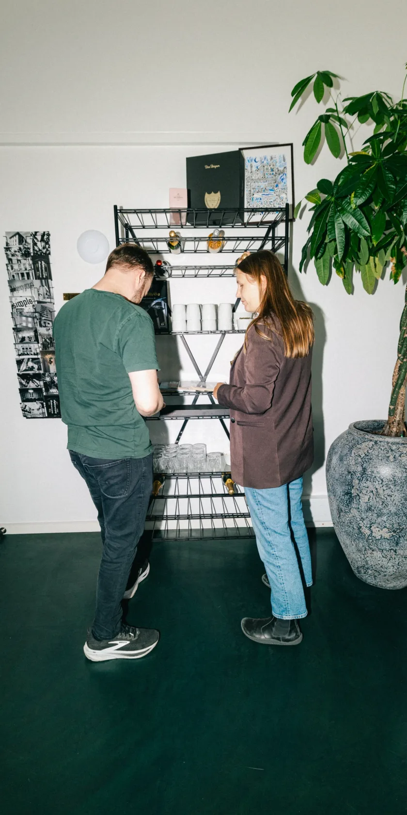 To kolleger står ved en trådreol med glas og flasker i et lyst lokale
