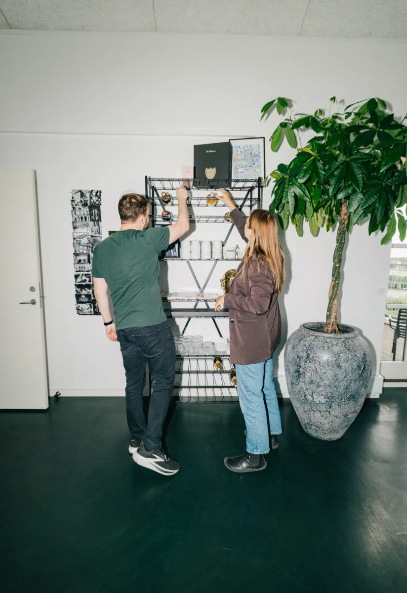 To kolleger rækker op efter noget på en trådreol ved en stor potteplante