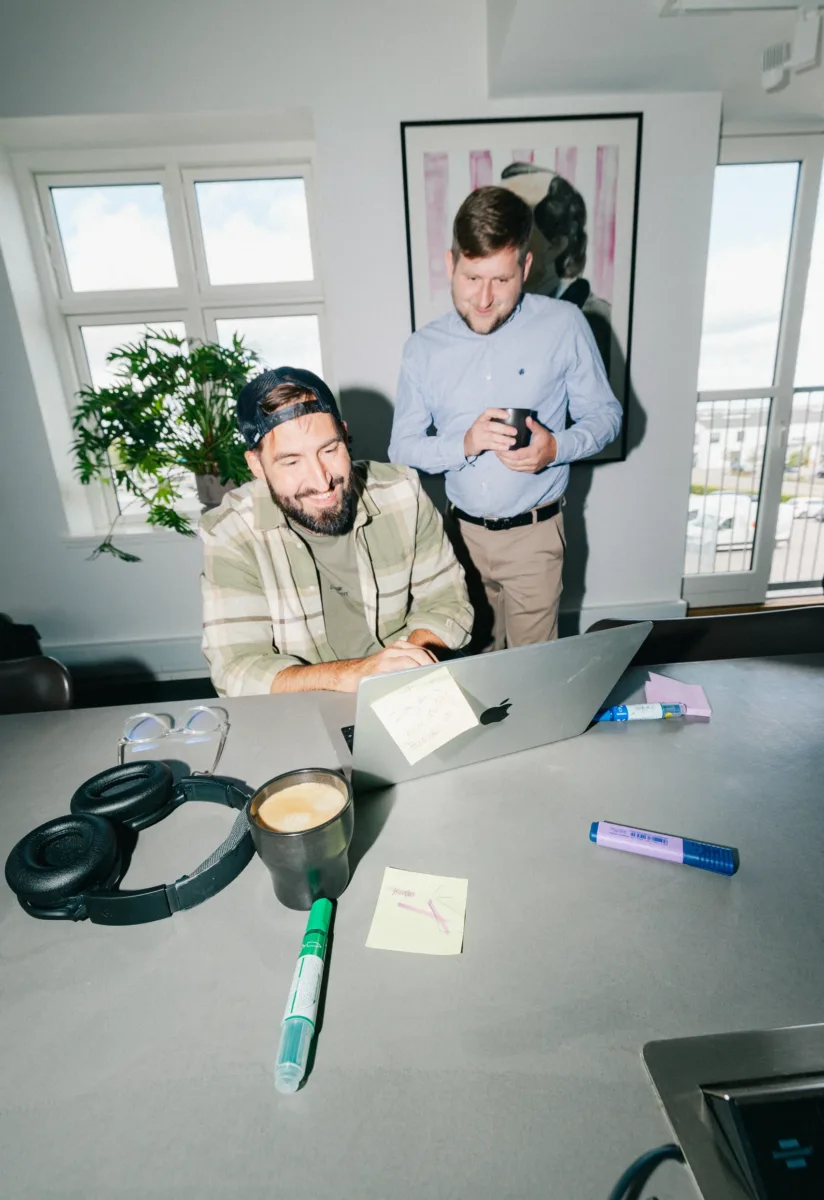 To kolleger smiler mod skærmen på en bærbar computer med noter på bordet