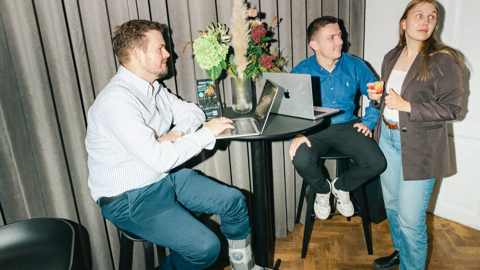 Kolleger taler sammen ved et højt bord med bærbare computere og tørrede blomster