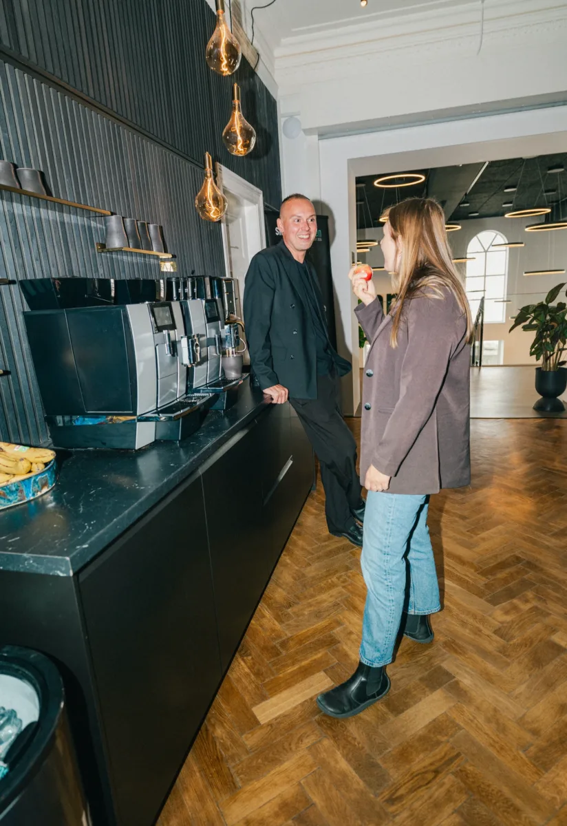 To kolleger snakker sammen ved kaffemaskinerne i et køkkenområde med sildebensgulv