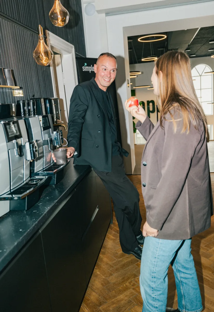 Kollega henter kaffe ved maskinen og smiler til en anden med et æble i hånden
